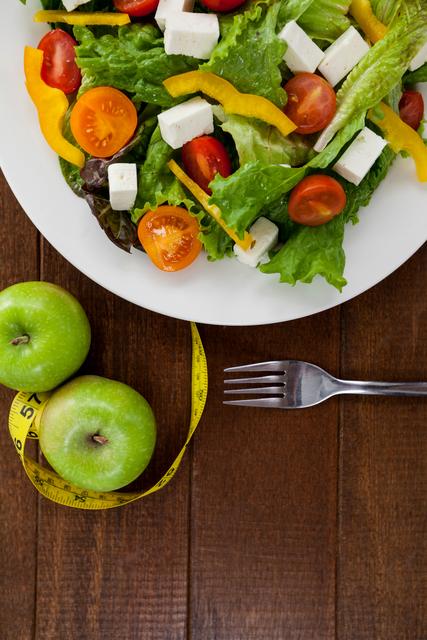 This image showcases a fresh and colorful salad with tomatoes, bell peppers, and feta cheese on a white plate, accompanied by green apples and a measuring tape on a wooden table. Ideal for use in health and wellness blogs, diet and nutrition articles, fitness programs, and healthy lifestyle promotions.