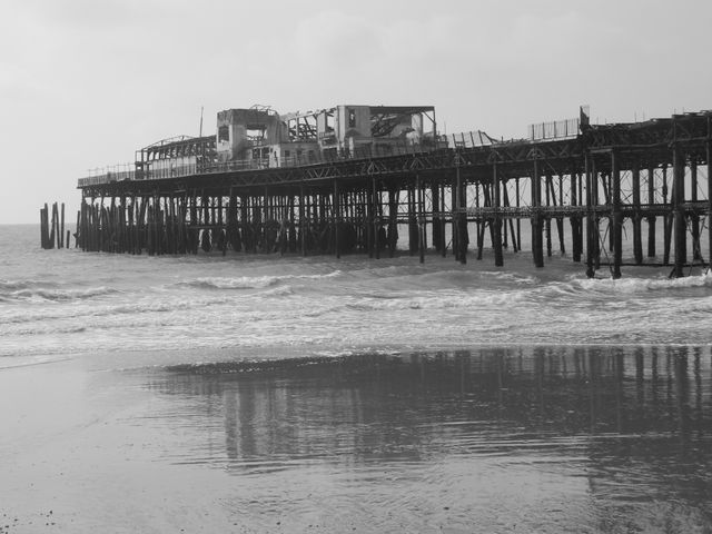 Old abandoned pier extending into ocean with shoreline and waves; reflecting in water on beach. Great for themes of nostalgia, decay, history, seaside, eerie, or solitude, suitable for travel websites, historic preservation content, or atmospheric artwork.