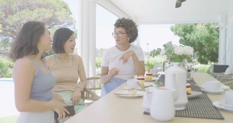 Three women chatting and sipping tea on covered patio by pool with cakes and flowers