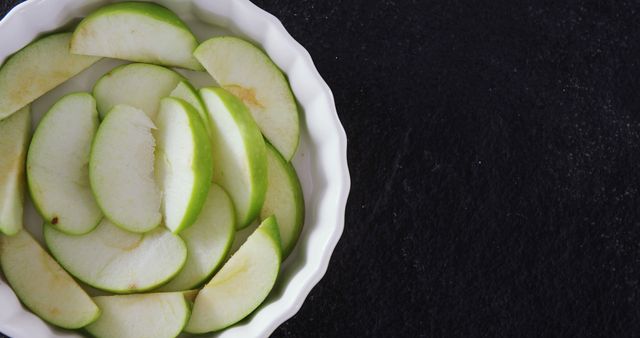 Sliced green apples are arranged neatly in a white bowl on a dark background, with copy space. Fresh apple slices like these are often used in healthy eating and culinary presentations.