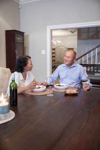 Asian senior couple sharing candlelit dinner at home, smiling over wine and chopsticks