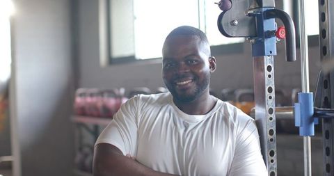 African american man standing confidently with arms crossed in gym near squat rack in white shirt