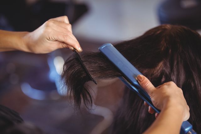 Hairdresser using a straightener and comb to style client's hair at a salon. Ideal for use in beauty and fashion blogs, hair care product advertisements, salon promotional materials, and hairstyling tutorials.