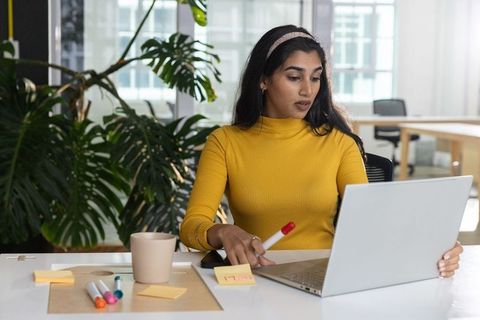 Indian woman working on laptop in modern open-plan office holding marker and sticky notes