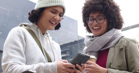 Smiling friends checking phone in downtown plaza holding coffee and wearing casual layers