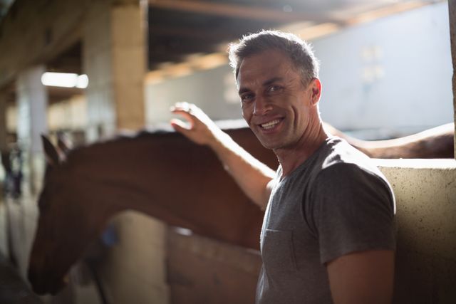 Smiling man caressing the brown horse in the stable