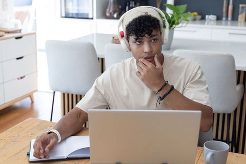 Young man working from home kitchen, thinking while using laptop and wearing headphones