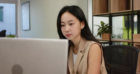 Focused asian woman working on laptop in modern office space
