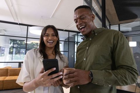 Smiling diverse coworkers sharing phone and coffee in modern office lounge teamwork