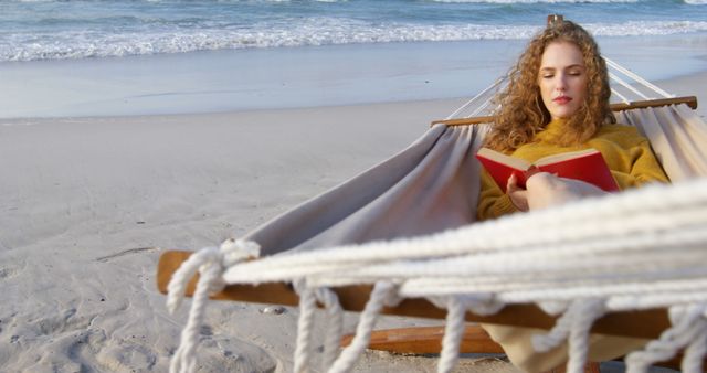 This photo depicts a woman enjoying a leisurely day in a hammock on a sandy beach, reading a book as she relaxes by the ocean. The sea waves in the background add to the tranquil and sunny atmosphere. Ideal for advertising vacation destinations, promoting relaxation products, or illustrating concepts related to reading, tranquility, and beach holidays.