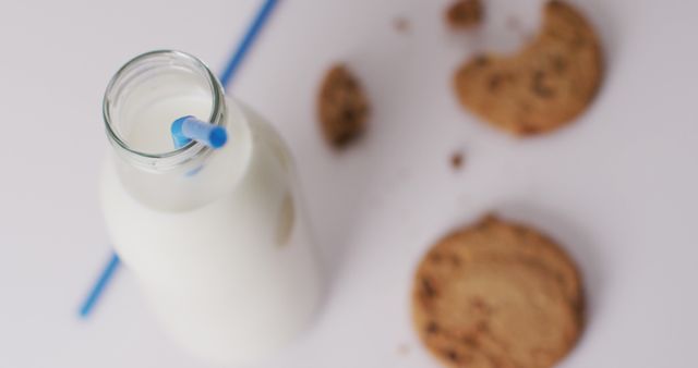 Glass bottle of milk and scattered chocolate chip cookies on a white surface. Perfect for illustrating snack time, dessert recipes, comforting food, and classic childhood memories.