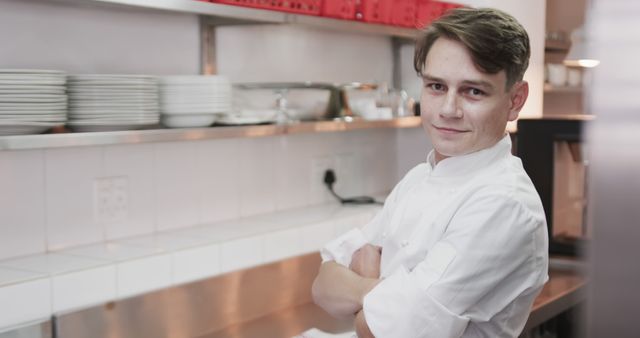 Young chef standing with folded arms in a well-equipped professional kitchen. He is wearing a white chef coat. Stainless steel counter and neatly stacked dishes are visible in the background, conveying a sense of order and professionalism. Ideal for use in content about culinary arts, restaurant job advertisements, professional kitchen setups, or cooking tutorials.