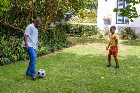 African american man playing soccer with boy on sunlit backyard lawn