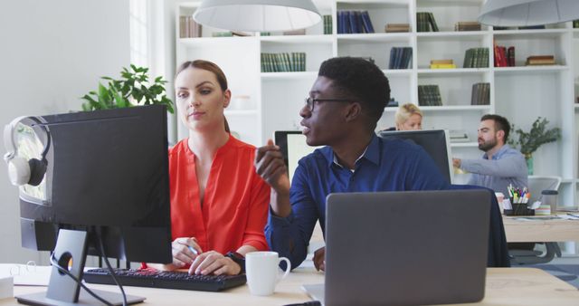 Two colleagues, one wearing a red shirt and the other a blue shirt, are having a discussion in front of a computer in a modern, well-lit office. Both are engaged and focused. This setting indicates teamwork and professional collaboration. Ideal for materials related to business environments, teamwork concepts, office settings, and professional workspaces.