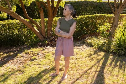 Senior asian woman standing in sunlit backyard wearing smartwatch and casual summer outfit