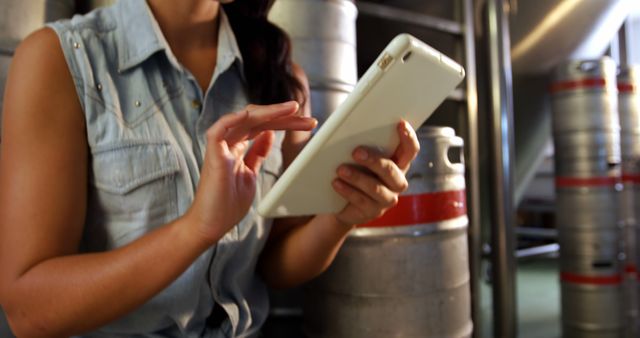 This stock photo features a woman using a tablet in a brewery setting. She appears focused and engaged while using the touchscreen device, with beer kegs and industrial equipment visible in the background. This image can be used for themes related to technology, business, modern industry, productivity, and casual work environments. Ideal for articles, blog posts, or marketing materials highlighting technology integration in traditional industries.