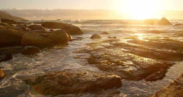Golden sunlight bathes a rocky shoreline at sunset, creating a serene outdoor scene. Waves gently lap against the stones, offering a moment of tranquility and natural beauty.