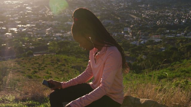 This image depicts a woman sitting outside, holding a smartphone, while wearing earbuds and exercising at sunset. The view of the countryside in the background highlights a combination of technology and nature. The photo is perfect for promoting products related to fitness, outdoor activities, or wearable technology, and can also be used in a lifestyle blog or wellness magazine focusing on exercise in natural environments.