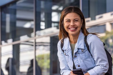 African american commuter smiling while holding smartphone by reflective glass facade