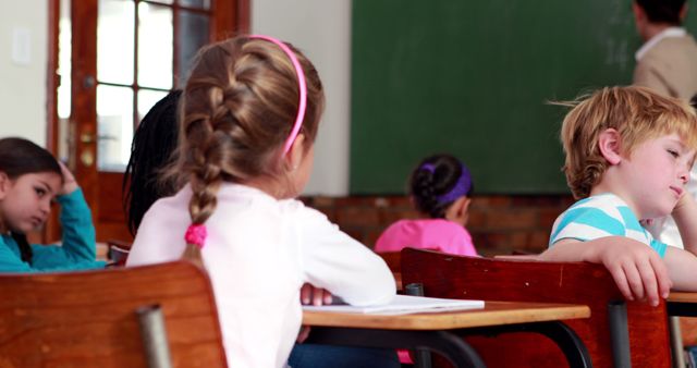 Children are sitting at desks in classroom, appearing bored and tired. Classroom setting with chalkboard and wooden desks suggests formal learning environment. Could be used for topics related to education challenges, student engagement, classroom management, and back-to-school content.