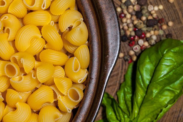 Uncooked pasta in a brown ceramic bowl surrounded by fresh basil leaves and mixed peppercorns on a wooden surface. Perfect for highlighting raw ingredients in cooking blogs, recipe websites, or Italian cuisine promotional materials. The vibrant colors and detailed close-up can draw attention to culinary skills and food photography.