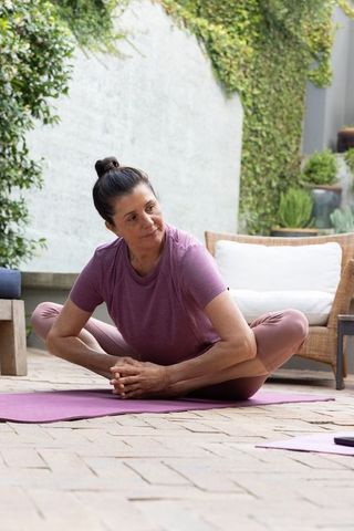 Mature woman practicing seated bound-angle yoga on patio mat in leafy courtyard wearing mauve leggin