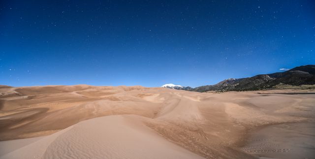 The image captures a tranquil scene of desert sand dunes under a starry night sky, with mountains in the background. It is excellent for use in nature magazines, travel brochures, and websites that focus on outdoor photography or remote natural landscapes. The serene setting can convey themes of peace, solitude, and natural beauty.