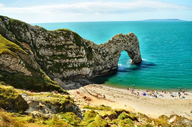 Beautiful view of Durdle Door, the iconic limestone arch on the Jurassic Coast, with tourists enjoying the beach and clear turquoise waters. This image is perfect for travel magazines, blogs showcasing natural wonders, coastal scenery travel guides, and vacation advertisements highlighting summer holiday destinations. The striking colors and stunning landscape make it an ideal visual for promoting outdoor adventures and scenic spots in Great Britain.