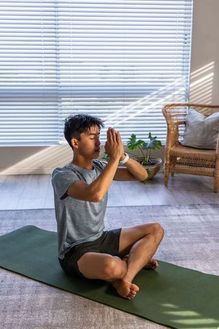 Asian man meditating on green yoga mat at home with blinds casting natural light