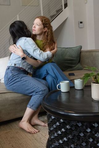 Women hugging on sofa in cozy living room, casual friendship moment with coffee, denim jacket