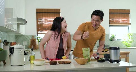 Laughing couple cooking together at modern kitchen island, squeezing lemon into blender