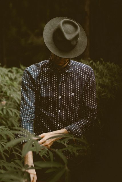 Mysterious man wearing a hat, standing among lush greenery, creating a sense of adventure and exploration. Ideal for use in travel promotions, outdoor activity advertisements, nature blogs, fashion lookbooks, and lifestyle magazine articles focusing on exploration and fashionable outdoor attire.