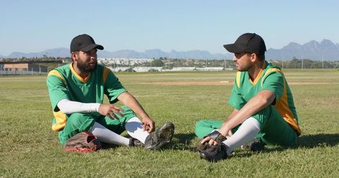 Multiracial baseball teammates sitting on grass talking and resting during sunny practice