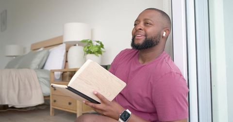 African man journaling by bedroom doorway wearing earbuds and smartwatch, sunlit minimal