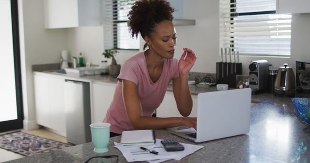 Young woman in modern kitchen working on laptop while reviewing documents and using calculator. Perfect for depicting themes related to remote work, freelancing, modern lifestyles, productivity, and professional women balancing home and work life.