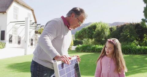 Grandfather teaching granddaughter about solar panel technology in sunny suburban garden