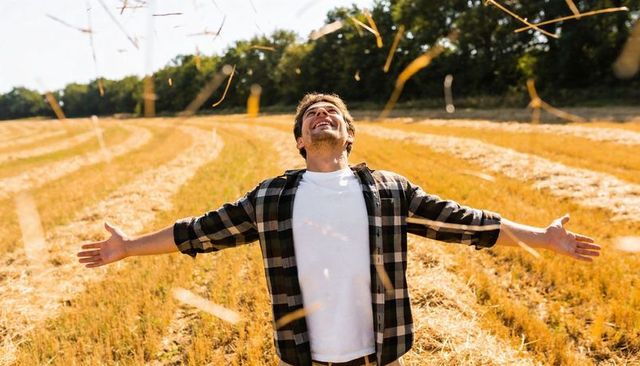 Joyful farmer spreading arms in golden harvested field under warm sunlight and floating straw