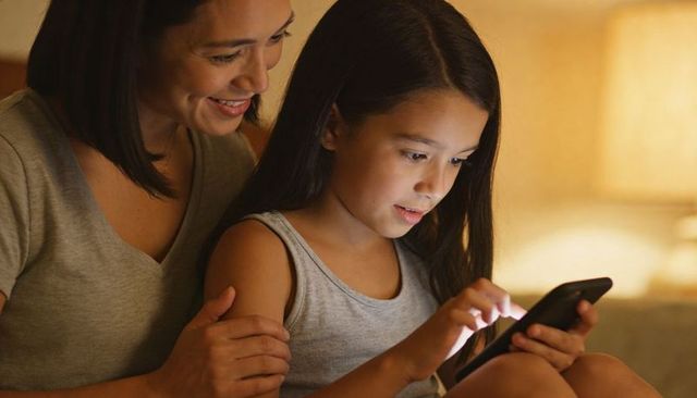 Smiling mother and daughter sharing cozy evening screen time on sofa with warm lamp glow