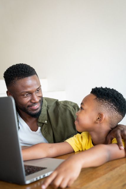 African american father and son sitting at desk and using laptop. family life, spending time together at home.