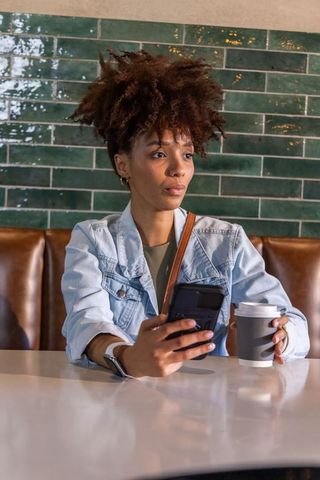 African american woman sitting at cafe holding smartphone and coffee cup in denim jacket