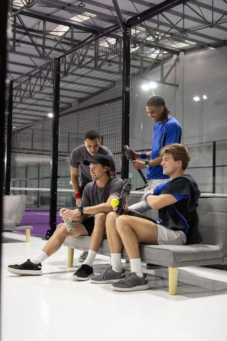 Group of male teammates resting and laughing at indoor padel court with paddles and ball