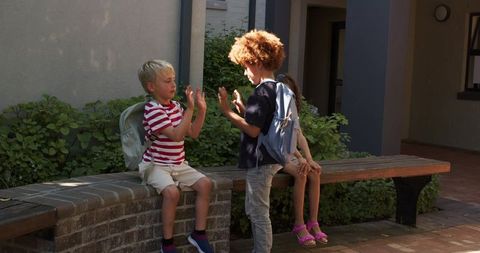 Children playing hand-clap game and laughing on sunlit courtyard bench with backpacks