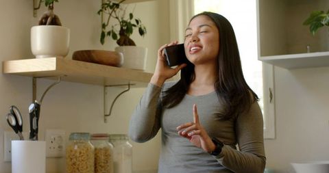 Asian woman chatting on smartphone in minimalist kitchen, smiling and gesturing casually