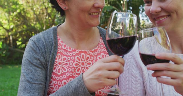 Two women seen toasting with glasses of red wine while enjoying an outdoor setting. Both are smiling happily and appear to be friends sharing a joyful moment. Ideal for use in marketing materials promoting friendship, outdoor gatherings, wine tours, and social celebrations.