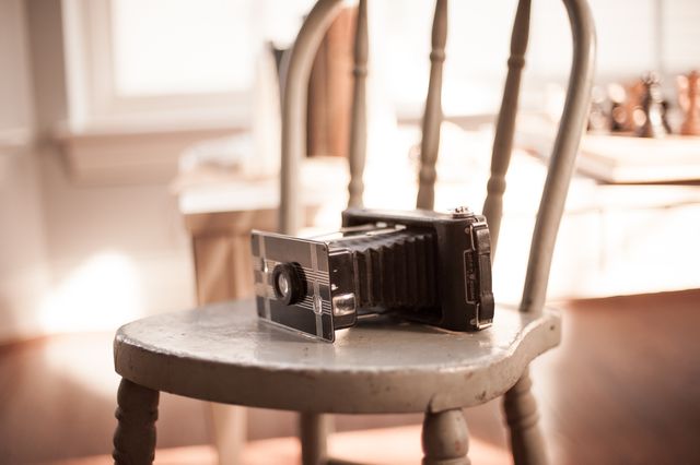 Still life displays vintage camera resting on timeworn wooden chair illuminated by natural sunlight. Ideal for use in historical photography, antique collections, decor, vintage lifestyle blogs.