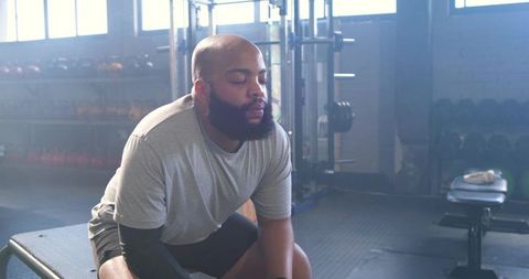 Bearded man sitting on flat bench resting after intense workout in sunlit weight room