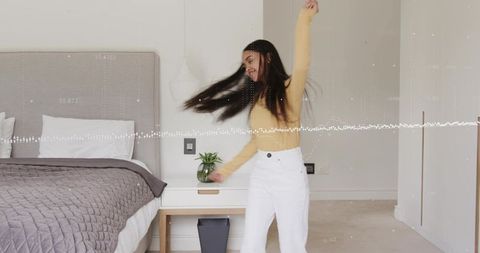 Young woman spinning in minimalist bedroom wearing mustard top and white high-waist pants