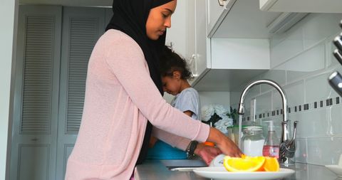 Young Woman in Hijab Washing Dishes with Daughter in Cozy Kitchen