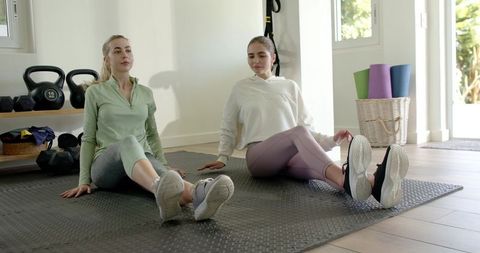 Two women stretching on mat in sunlit home gym with kettlebells and trx
