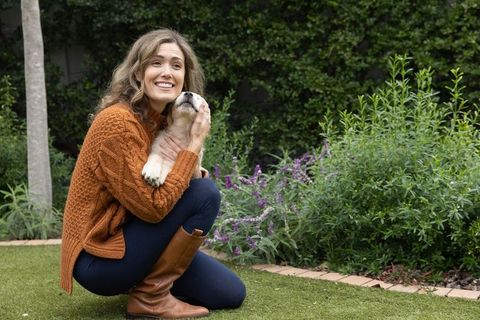 Smiling woman crouching in backyard cuddling light-colored puppy in cozy rust sweater